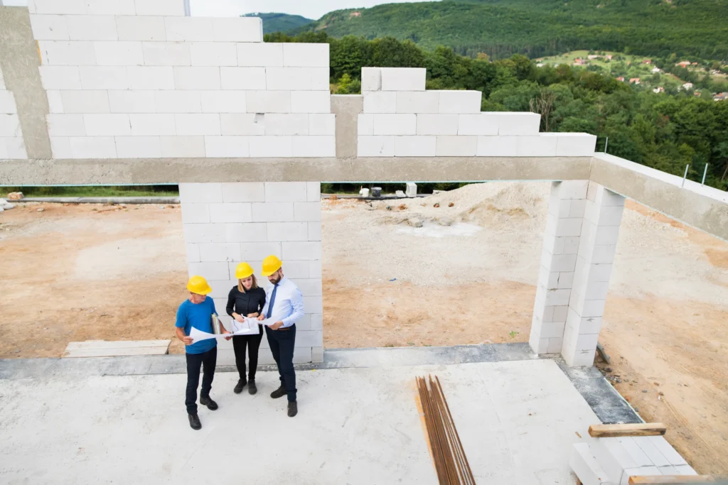 Three construction workers in yellow helmets review blueprints at a building site with partially constructed walls, surrounded by greenery and hills.