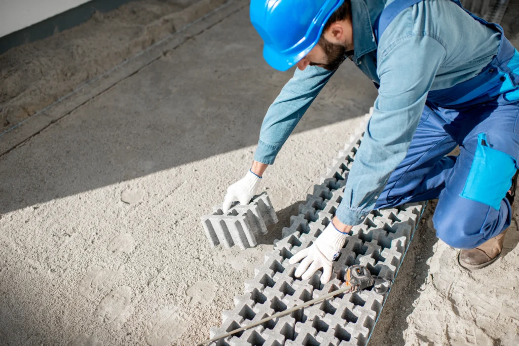 Worker in blue uniform and helmet lays concrete pavers on sand. The scene conveys construction, focus, and manual labor in progress.