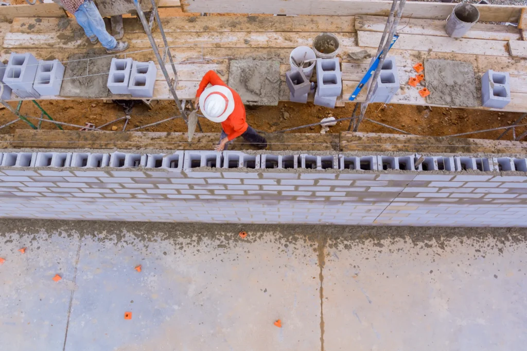A construction worker in a white hard hat and orange shirt lays cement blocks on a wall, viewed from above. Tools and materials are scattered around.
