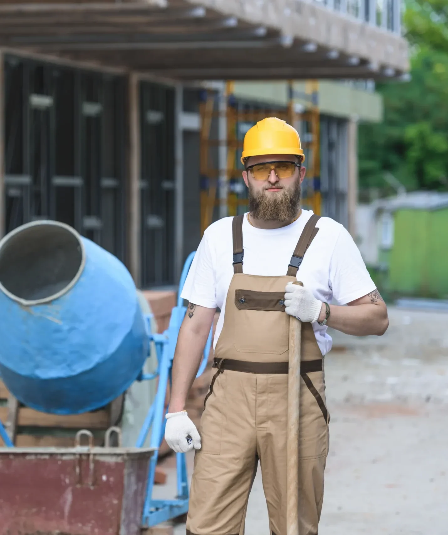 A construction worker in a yellow hard hat and brown overalls stands confidently holding a shovel. A blue cement mixer is nearby on a construction site.